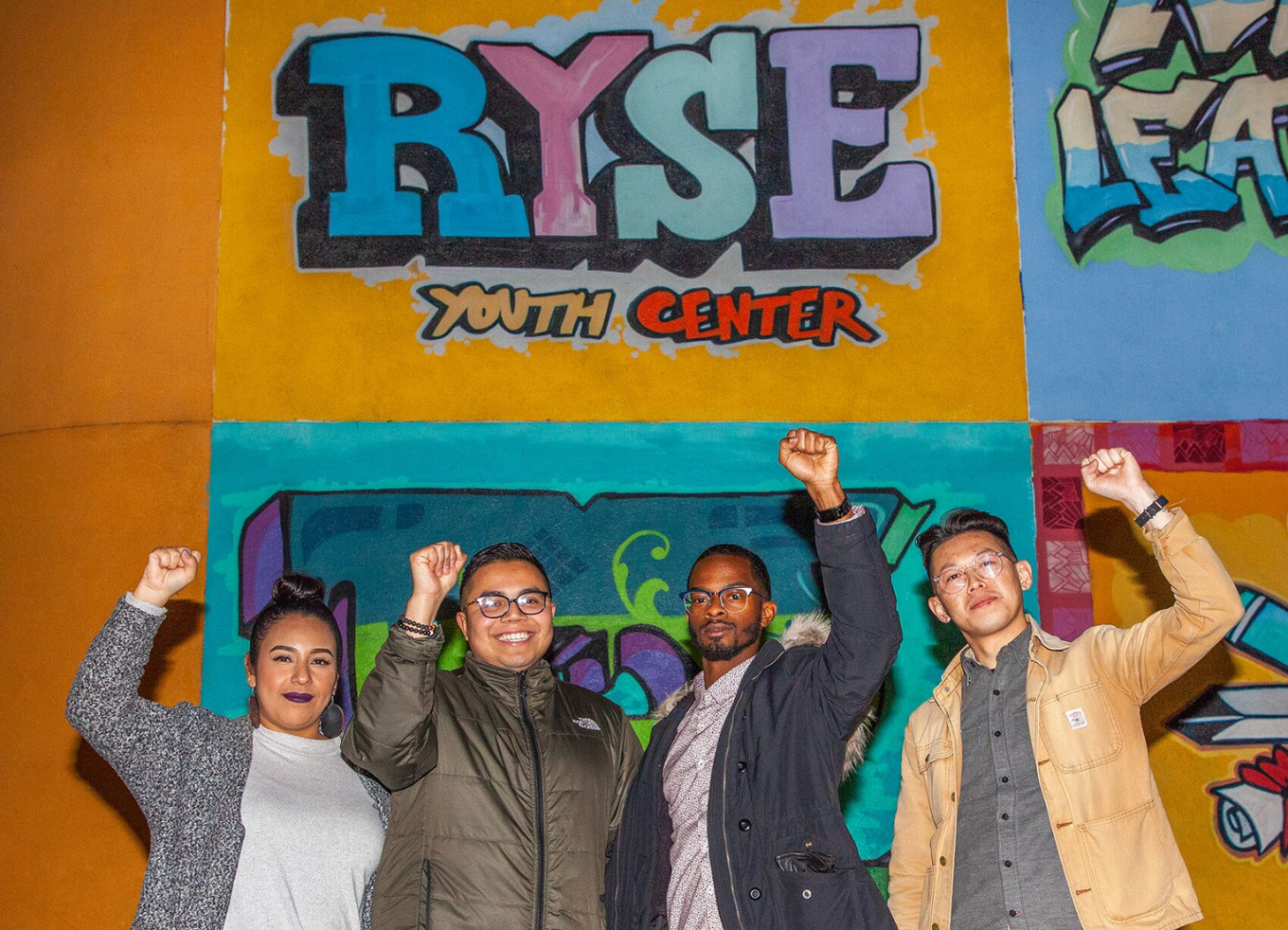 Four people stand with fists raised in front of a mural at the RYSE Youth Center.