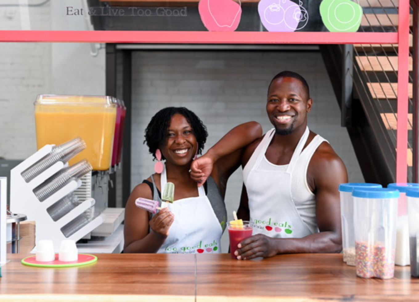 Two people behind a counter smiling while holding popsicles and a smoothie