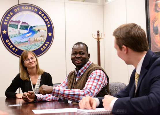 Man in sweater sitting at a desk with a man and woman wearing suits with the South Dakota state seal in the background.