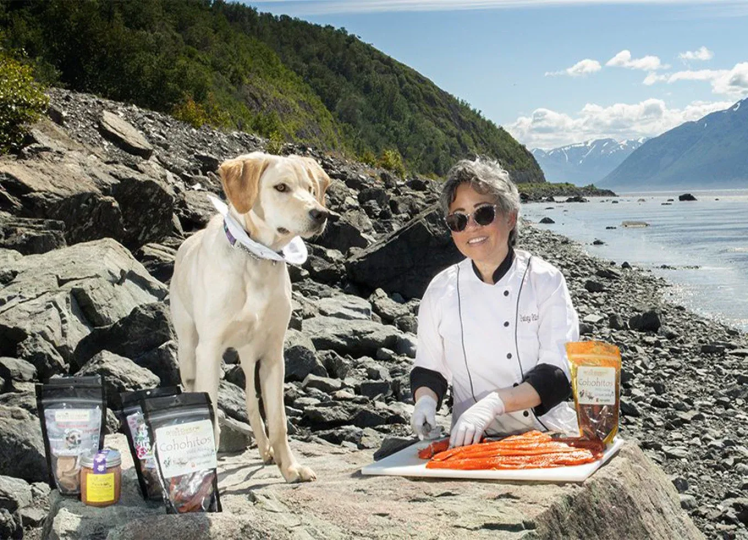 Daisy Nicolas, owner of Drool Central: A Mum and Pup Barkery, smiling and cutting fish with a dog next to her on a rocky shore.
